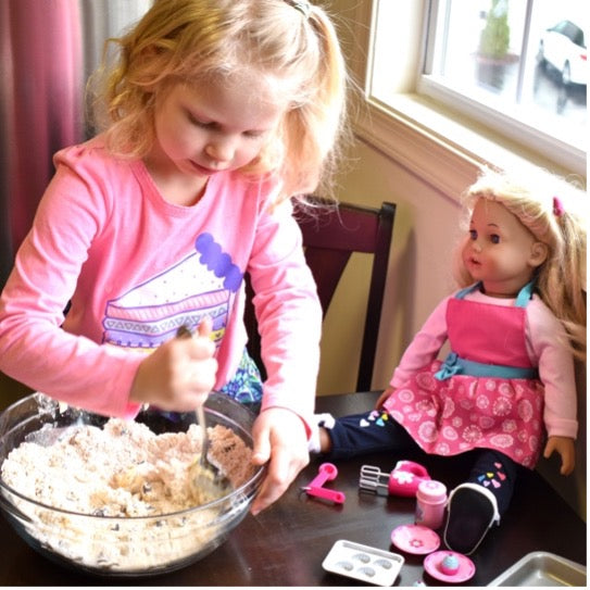 A little girl making cookie dough with her doll nearby with similar baking utensils