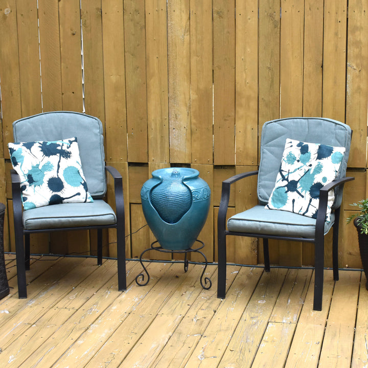 A teal vase-shaped water fountain between a pair of outdoor chairs on a patio