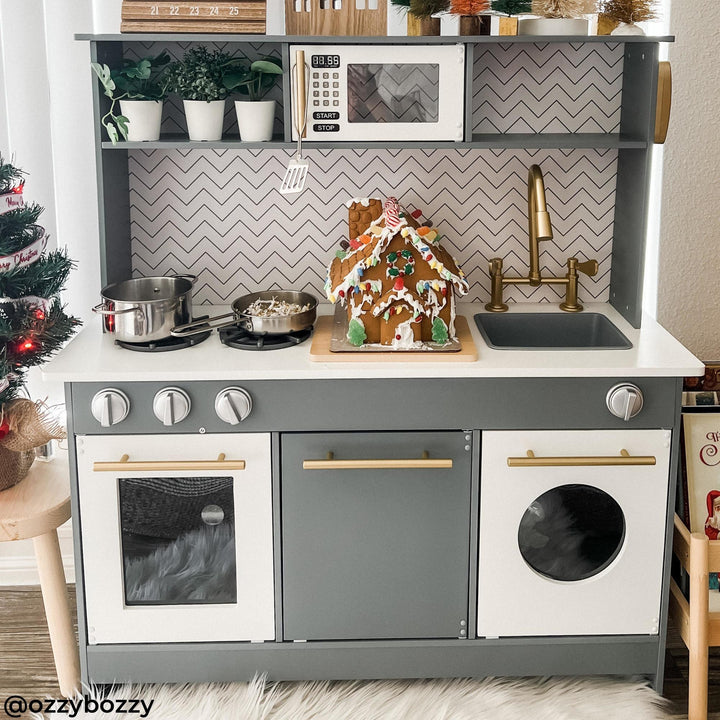 A gray and white play kitchen with a gingerbread house on the counter