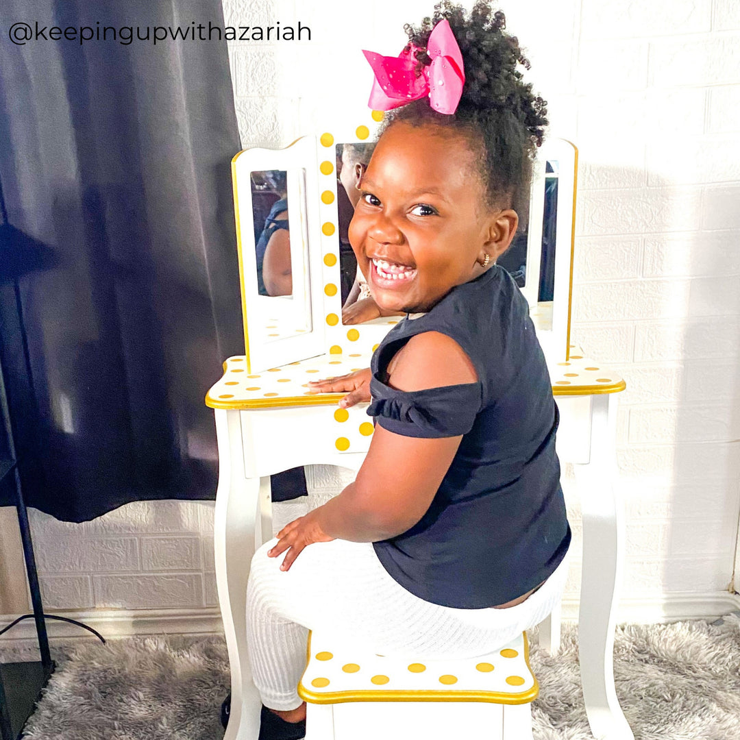 A little girl sitting at a white with gold polka dots vanity table 