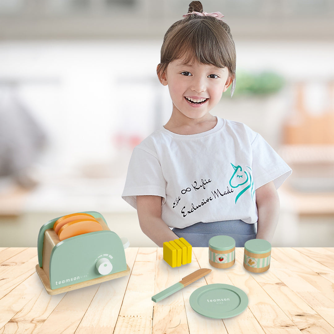 A little girl standing behing a table with a play toaster set spread out on the tabletop