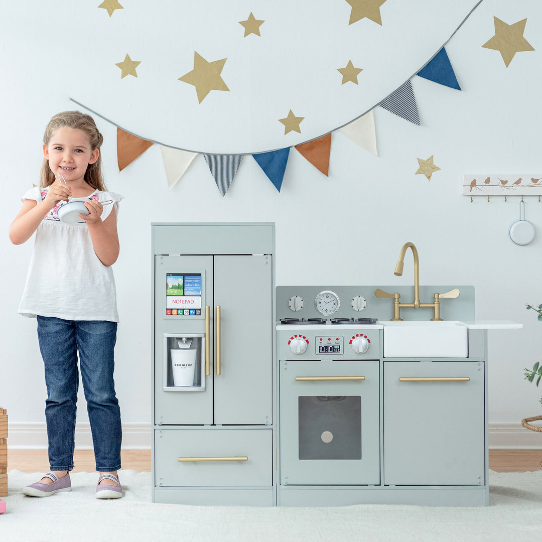 A young girl playing with a Teamson Kids Little Chef Charlotte Modern Play Kitchen, Silver Gray/Gold in a decorated room.