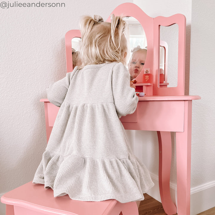 A little girl sitting at a pink vanity table looking in the mirror