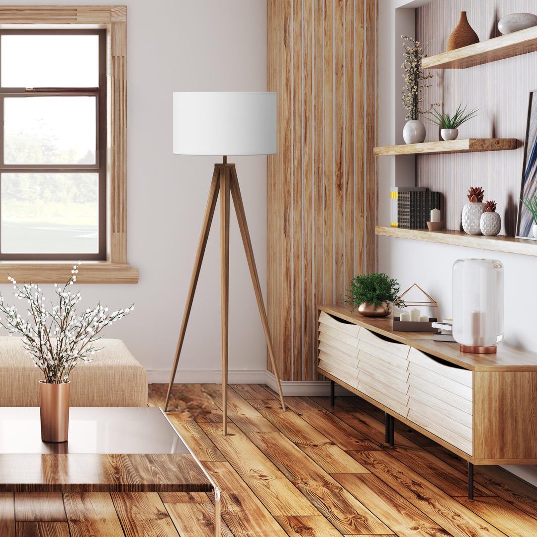 A tripod style floor lamp with natural wood finished legs and a white faux linen lampshade in a mid-century style living room