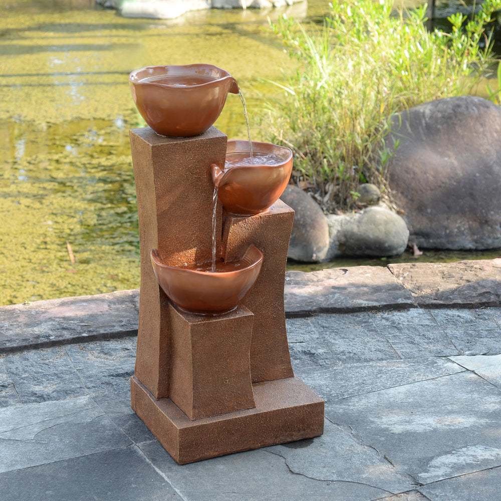 A view of a 3-Tier Outdoor Water Fountain with bowls stacked on a pedestal, with water flowing from top to bottom, located beside a pond.