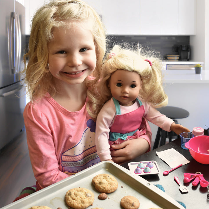 a young child smiles with her doll at a table with freshly baked cookies