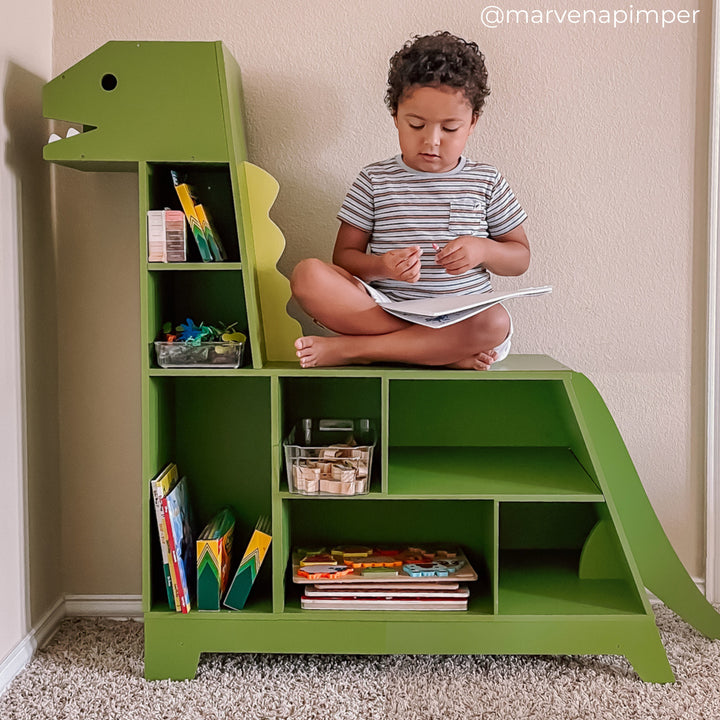 Child sits cross-legged and focused, writing on a notepad atop a green dinosaur-shaped bookshelf filled with toys and books.