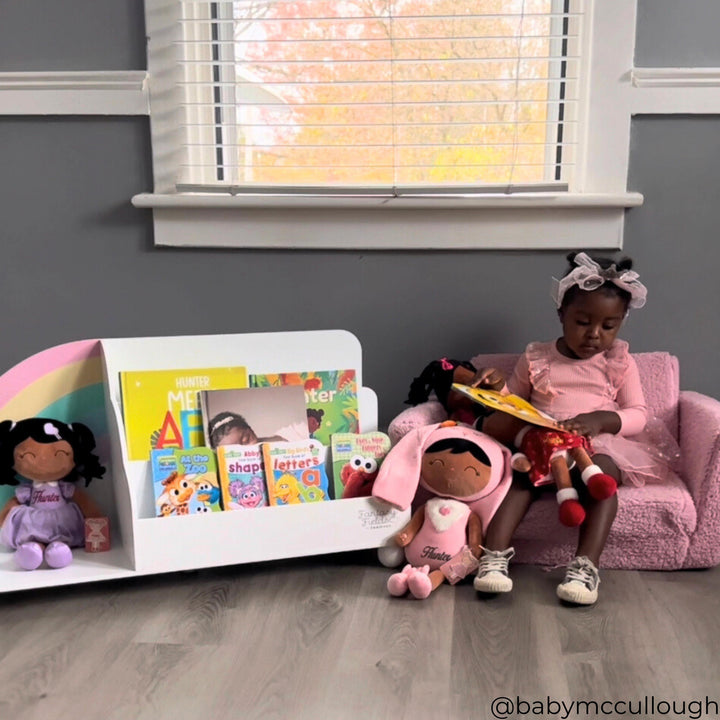 A little girl sitting on a pink chair looking at a book next to a short bookshelf