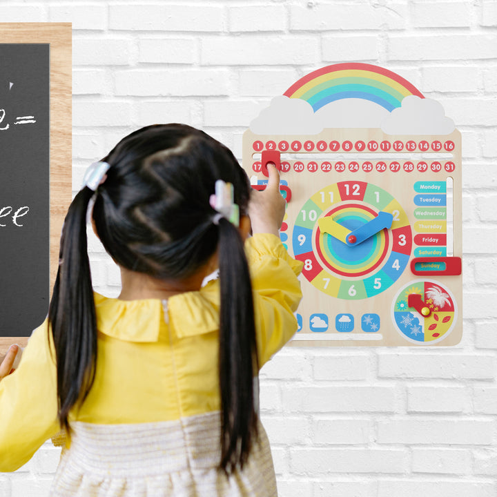 A little girl in a yellow and white dress uses her pointer finger to change the date number on the toddler daily calendar board, hanging on a wall