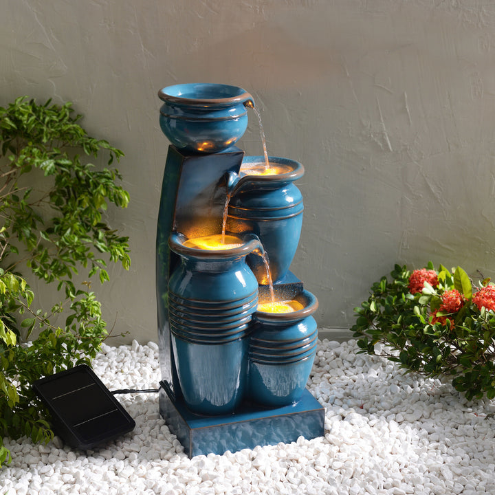 Teamson Home 28.74" Blue 4-Tier Outdoor Solar Water Fountain with LED Lights, surrounded by decorative plants and white pebbles.