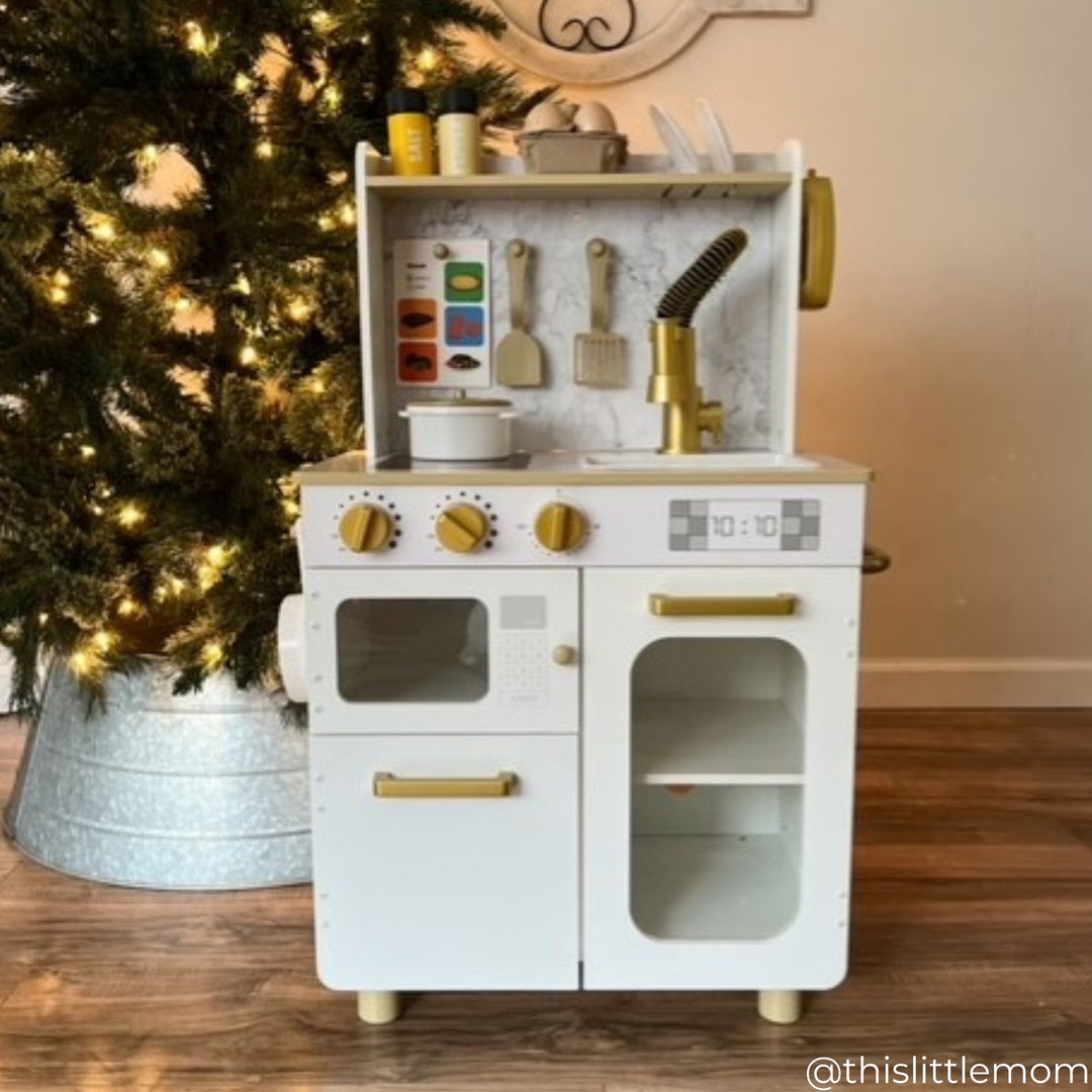 A white play kitchen with gold hardware next to a Christmas tree