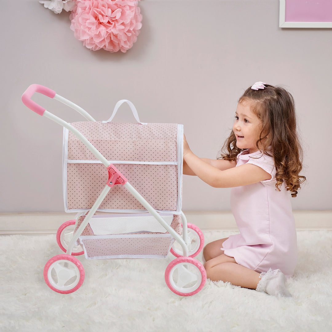 A young girl with a pink bow in her hair kneels on a white rug, playing with a pink and white toy stroller with polka-dotted fabric. Decorative pink pom-poms hang on the wall behind her.