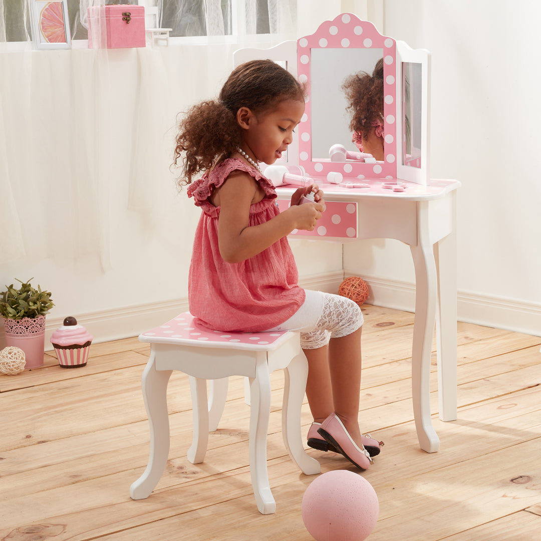 A little girl sitting on the matching stool next to her white vanity with pink and white polka dot accents