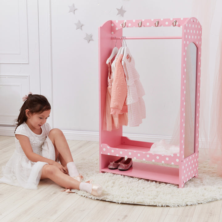 A girl sitting next to a Fantasy Fields Kids Polka Dots Clothing Rack with Storage