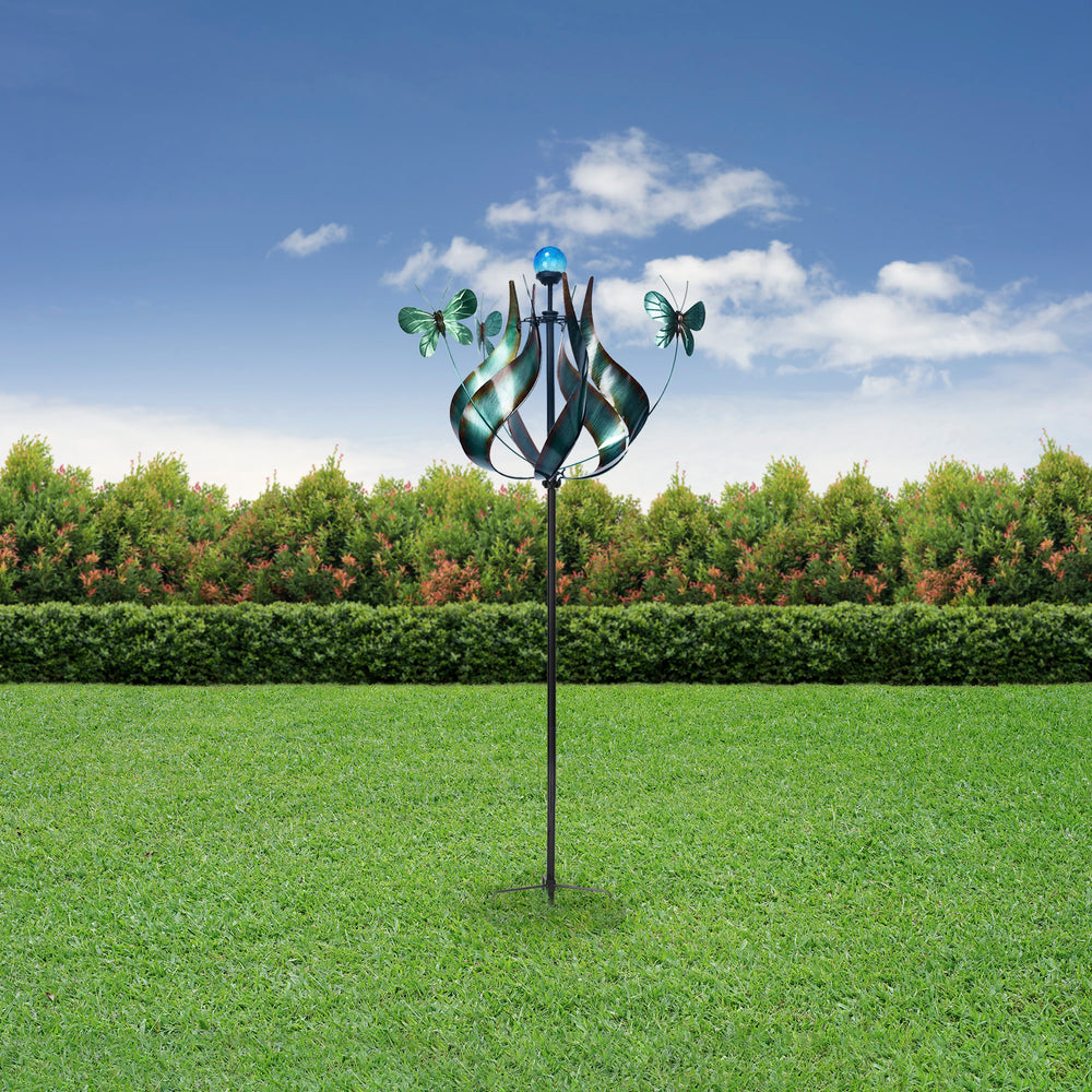 A colorful, Teamson Home Outdoor Solar Tulip and Butterfly Kinetic Windmill Sculpture with LED Light, Teal installed in a green lawn against a backdrop of hedges and a clear blue sky.