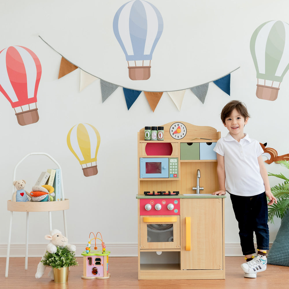 Boy smiling and standing next to Teamson Kids Little Chef Florence Classic Play Kitchen with 5 Kitchen Accessory Toys, Wood Grain