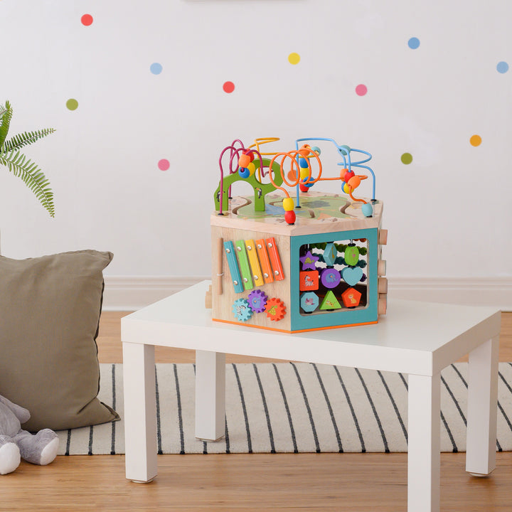 Colorful Teamson Kids Preschool Play Lab 7-in-1 Large Wooden Activity Station with bead mazes, gears, and shape sorters for children's learning and pla on a small white table in a playroom.