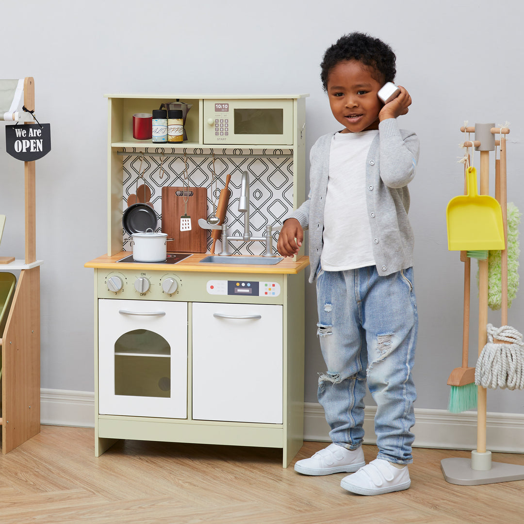 A little boy making a pretend phone call next to his Little Chef Boston Farmhouse Wooden Kitchen Playset, Green/White