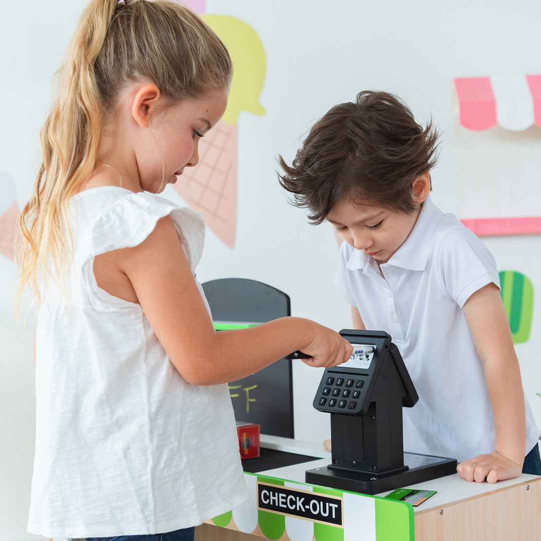 Two kids playing market stand with one pretending to check out the other's purchase at the Teamson Kids Cashier Austin Play Market Checkout Counter with 26 Accessories, Green/Natural.