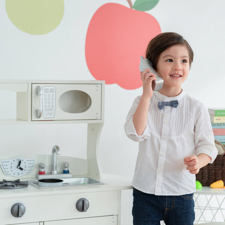 A young child pretends to talk on a Teamson Kids Little Chef Westchester Retro Kids Kitchen Playset, Ivory while playing with an interactive play kitchen set.