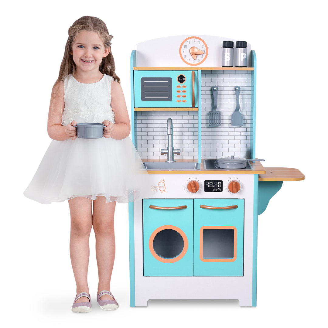 A smiling young girl standing next to a Teamson Kids Little Chef Santos Retro Wooden Kitchen Playset in Aqua/White with interactive features.