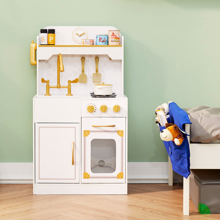 A play kitchen set in white with a faux marble backsplash and gold accents against a green wall next to a bed.