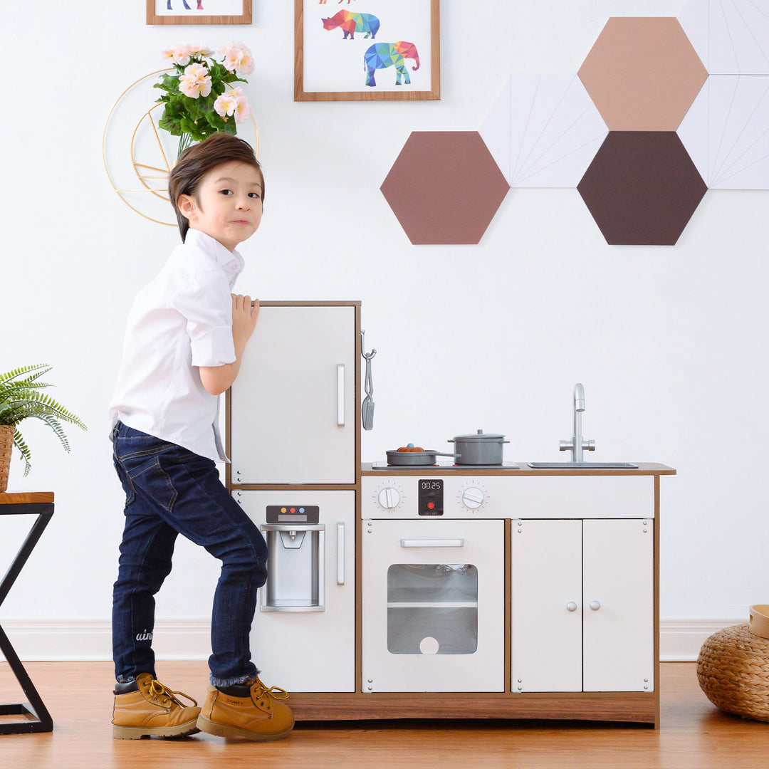 A child standing next to a Teamson Kids Little Chef Palm Springs Classic Kids Play Kitchen with Accessories, Natural/White, looking at the camera with a slight smile.