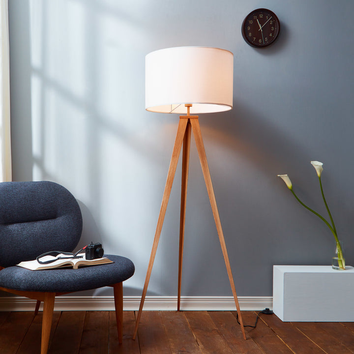 A tripod style floor lamp with natural wood finished legs and a white faux linen lampshade