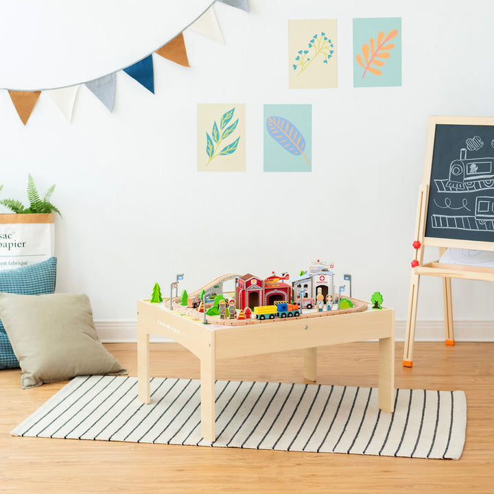 A well-organized children's play area with a Teamson Kids Preschool Play Lab Toys Wooden Table with 85-pc Train and Town Set, Natural on a table, including an intricate track arrangement, and an easel in a bright room.